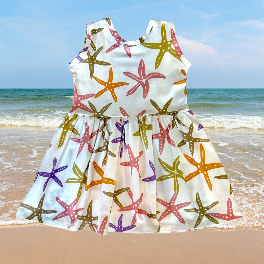 A sleeveless, organic cotton beach dress for children with a starfish pattern, displayed on a beach setting.