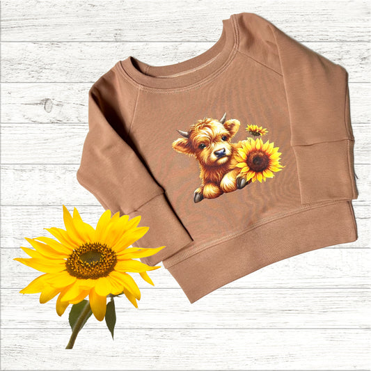A brown crew neck sweatshirt with a graphic print of a cow and sunflowers, displayed next to an actual sunflower.