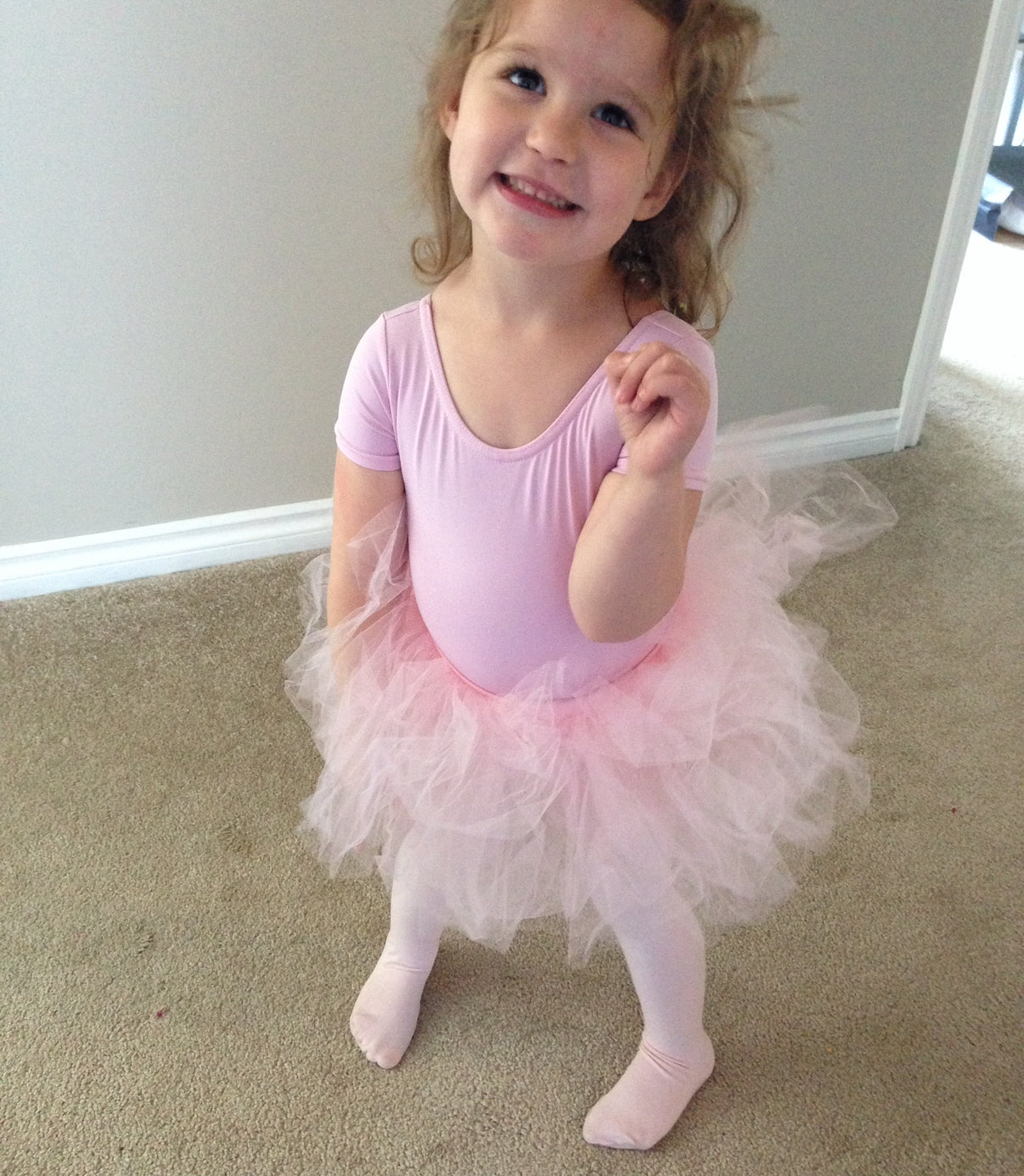 Young girl in a pink ballet outfit standing on a carpeted floor.